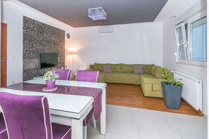 A modern living room with a dining area, a green sofa, a large potted plant, and a flat-screen TV mounted on a textured wall. - Apartment Mario (Strozanac)
