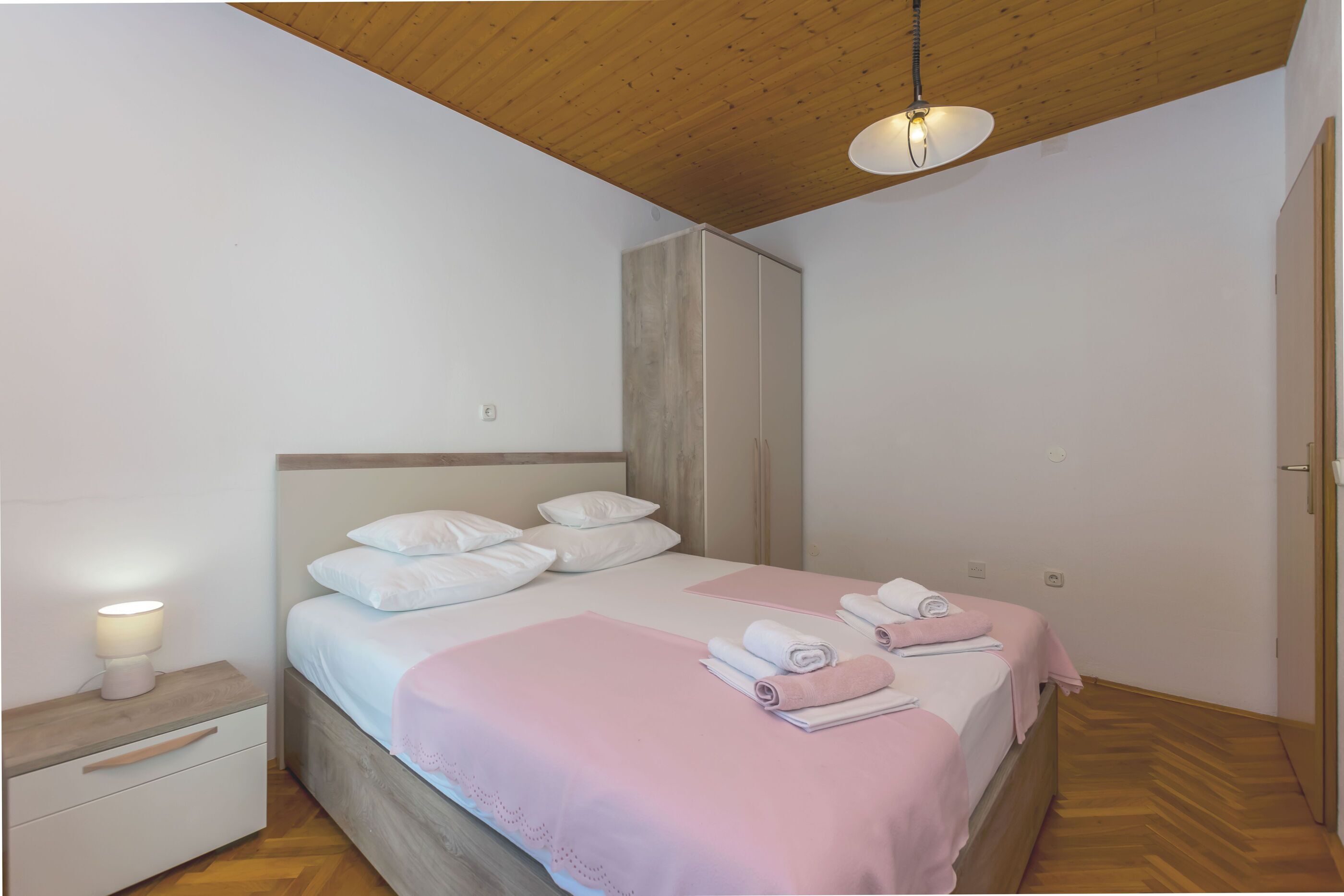 A bedroom with a wooden ceiling, a bed with white bedding, a bedside table with a lamp, and a wardrobe.