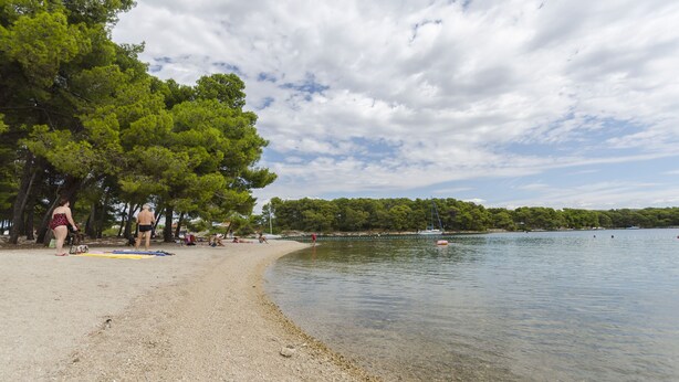 A pebbled beach with clear water, trees, and a partly cloudy sky. - Apartments Romic (Jadrija)