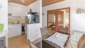 A compact kitchen with white cabinets, a built-in stove, and a refrigerator. There is a dining table with chairs and a glass top. - Apartments Romic (Jadrija)
