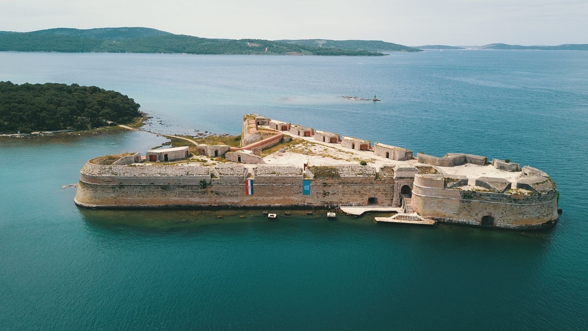 Eine Vogelperspektive zeigt eine historische Festung, umgeben von Wasser, eine kleine Insel und ein Schiff in der Ferne.