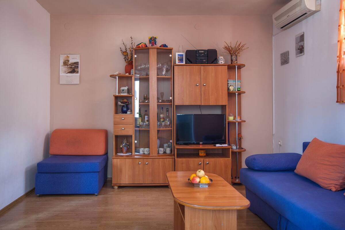 Un salon avec un canapé bleu, un fauteuil bleu, un meuble en bois doté de portes en verre, une télévision et une petite table sur laquelle se trouve des fruits.