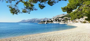 A pebble beach with a clear blue sea, a town with buildings on a hillside, and mountains in the background.