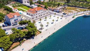 Ein Luftbild einer Küstenstadt mit einem markanten weißen Gebäude mit rotem Dach, einer gepflasterten Uferpromenade mit Palmen und einem klaren, blauen Meer.
