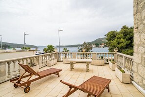 A terrace with wooden lounge chairs, a table, and potted plants, overlooking a body of water and hills.