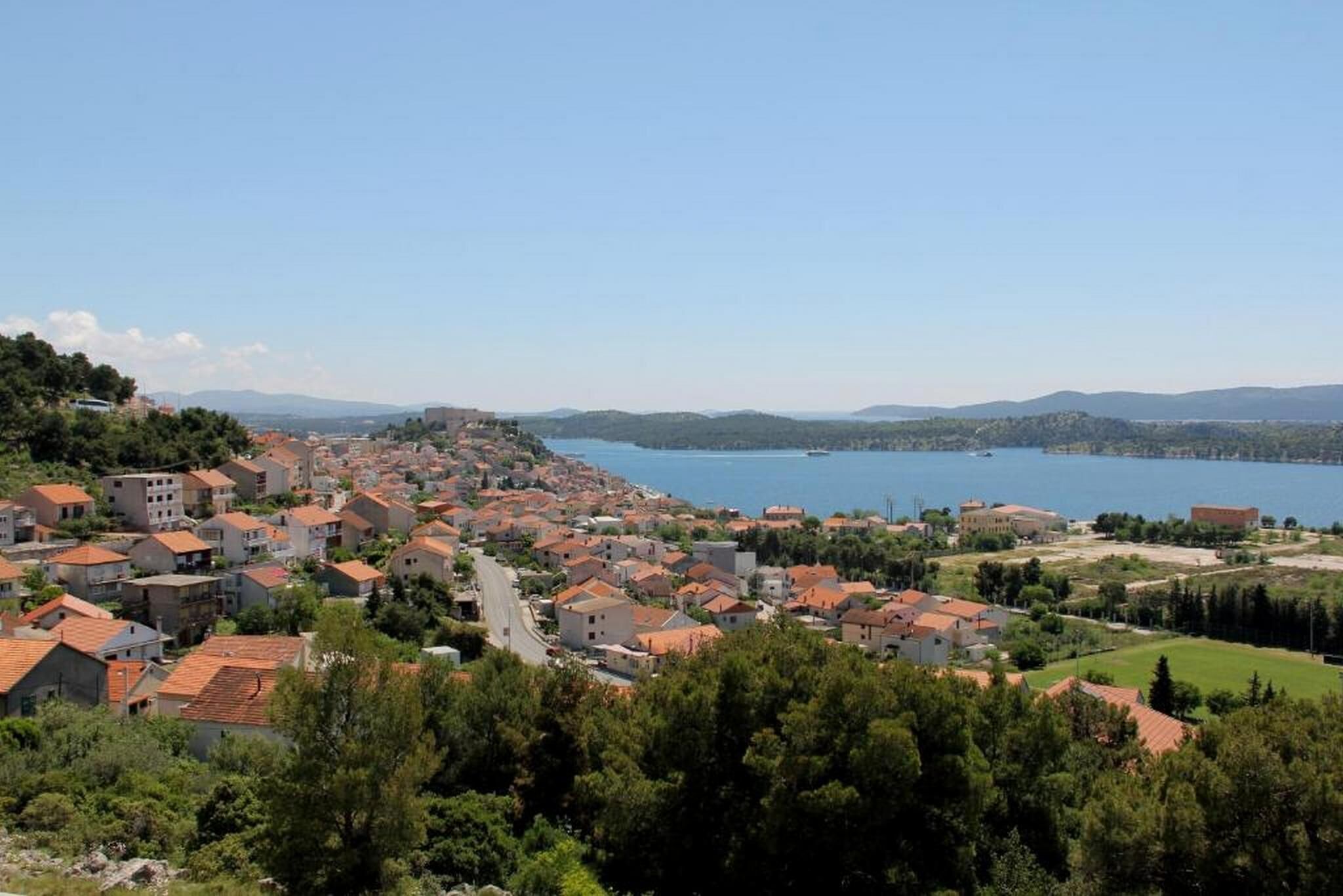 A coastal town with red-roofed buildings, a clear blue lake, and hilly terrain in the background.
