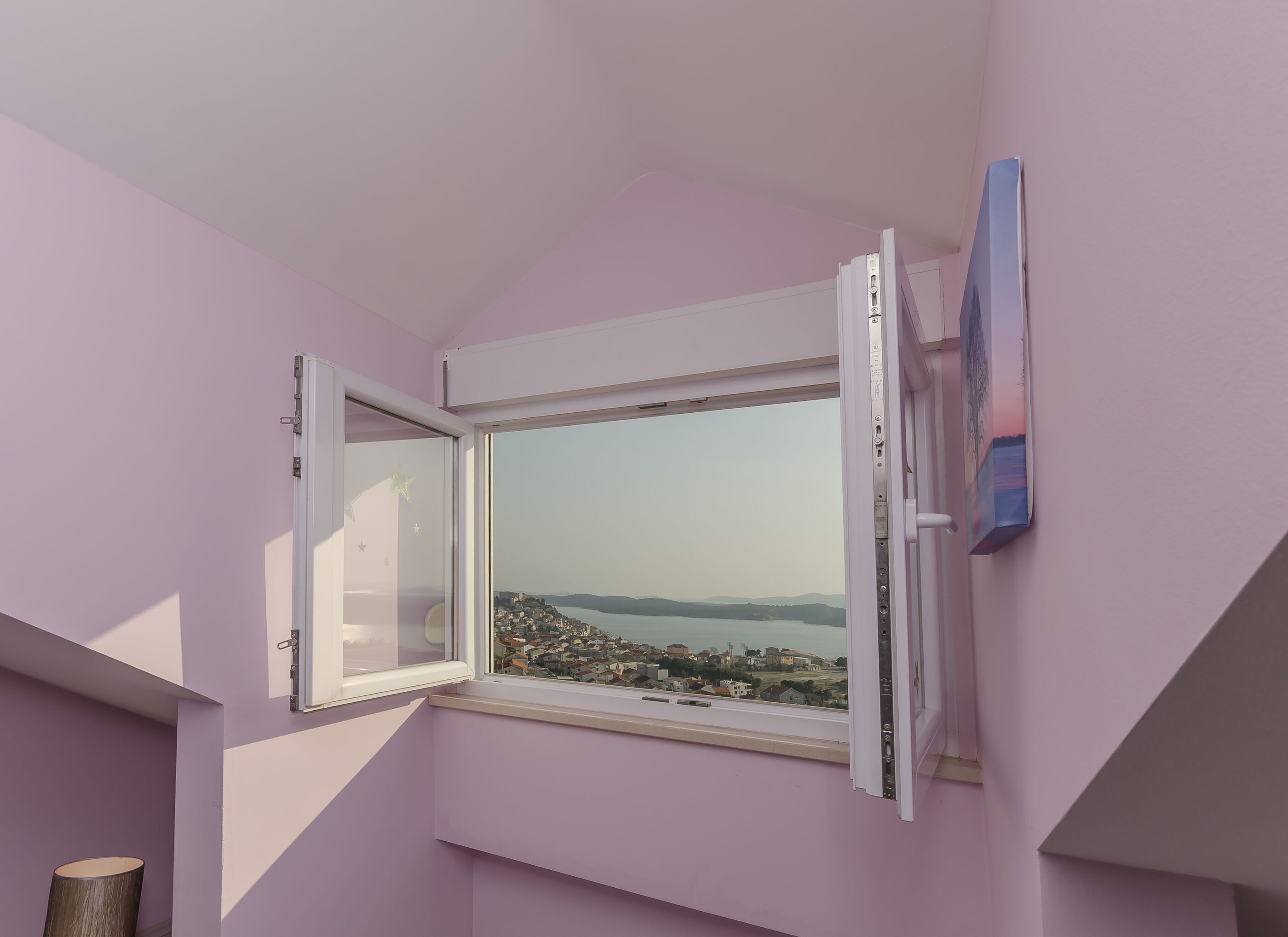 Ein Zimmer mit einer rosafarbenen Wand, einem Fenster mit Blick auf eine Stadt und Wasser, sowie einer Tür mit Griff.