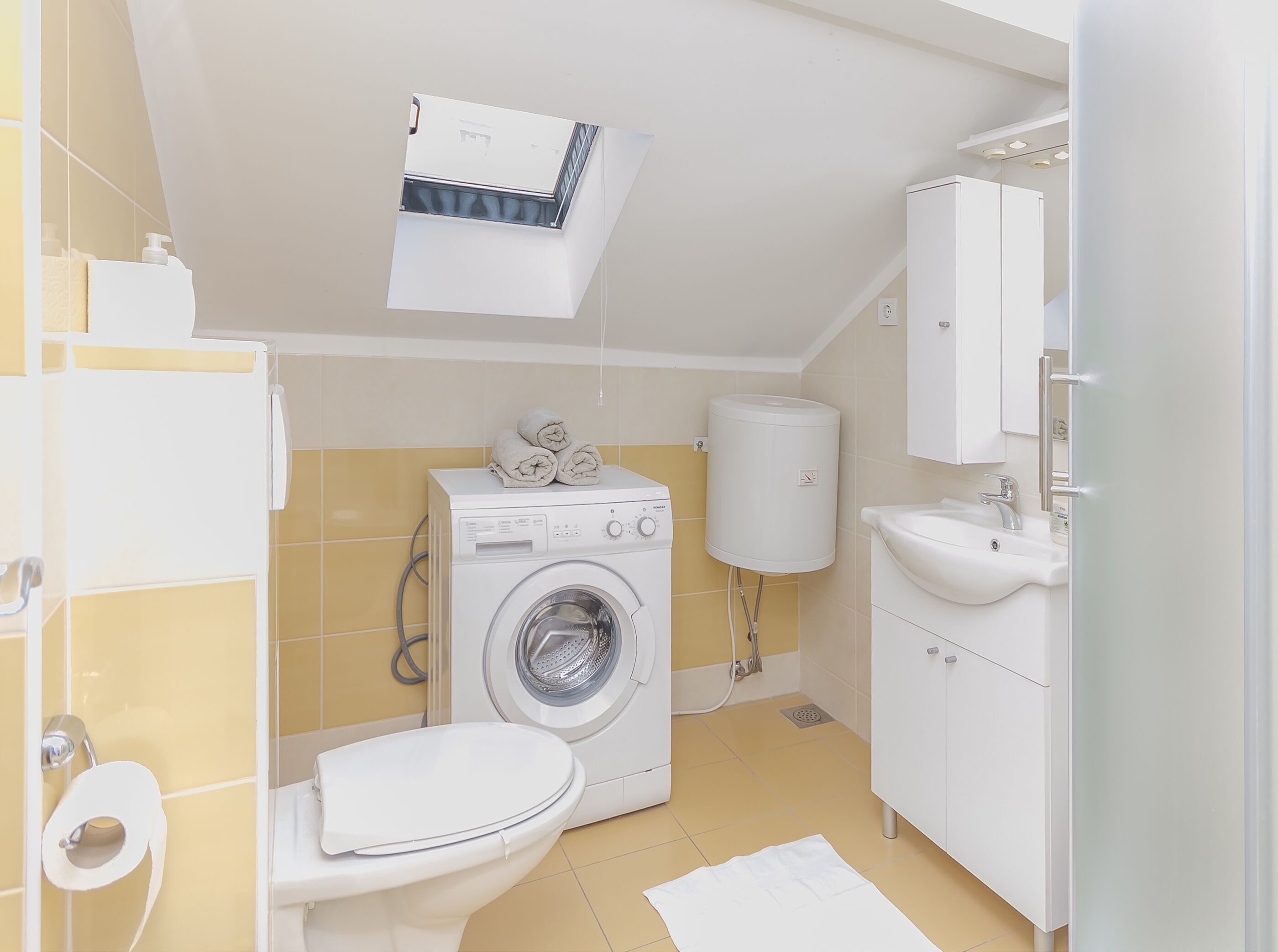 Een kleine badkamer met een wasmachine, toilet, wastafel en een klein raam boven de wasmachine.