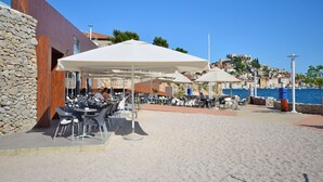 An outdoor café with tables and chairs under umbrellas, a stone wall, and a view of the sea and buildings in the background. - Apartment Golden Sunset (Sibenik)