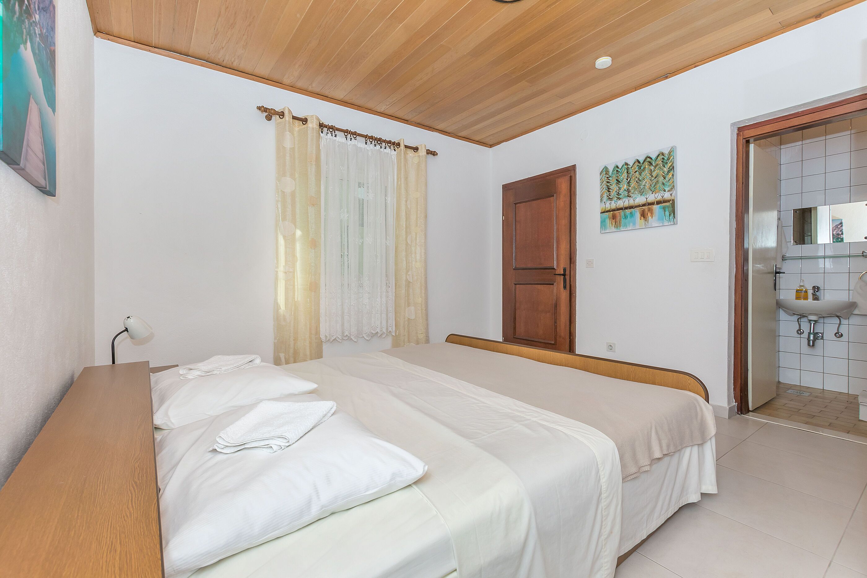 A bedroom with a wooden bed, a bedside table, a window with curtains, a door, and a bathroom with a sink and mirror.