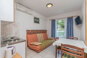 Eine kompakte Küchenzeile mit Spüle und Herd, ein Schlafsofa mit gemustertem Überwurf, ein Esstisch mit Stühlen und Blick ins Grüne durch das Fenster.