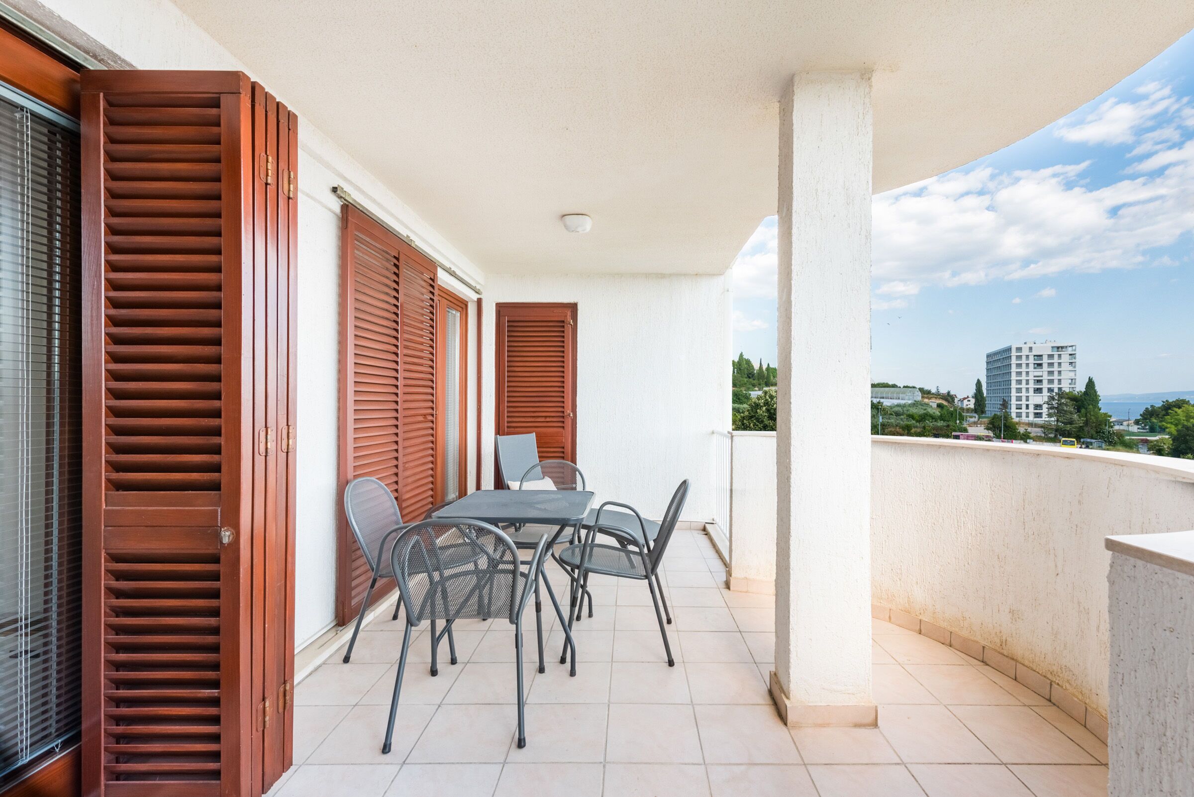 Un balcón con mesa y sillas, contraventanas de madera y vistas a edificios en la distancia.