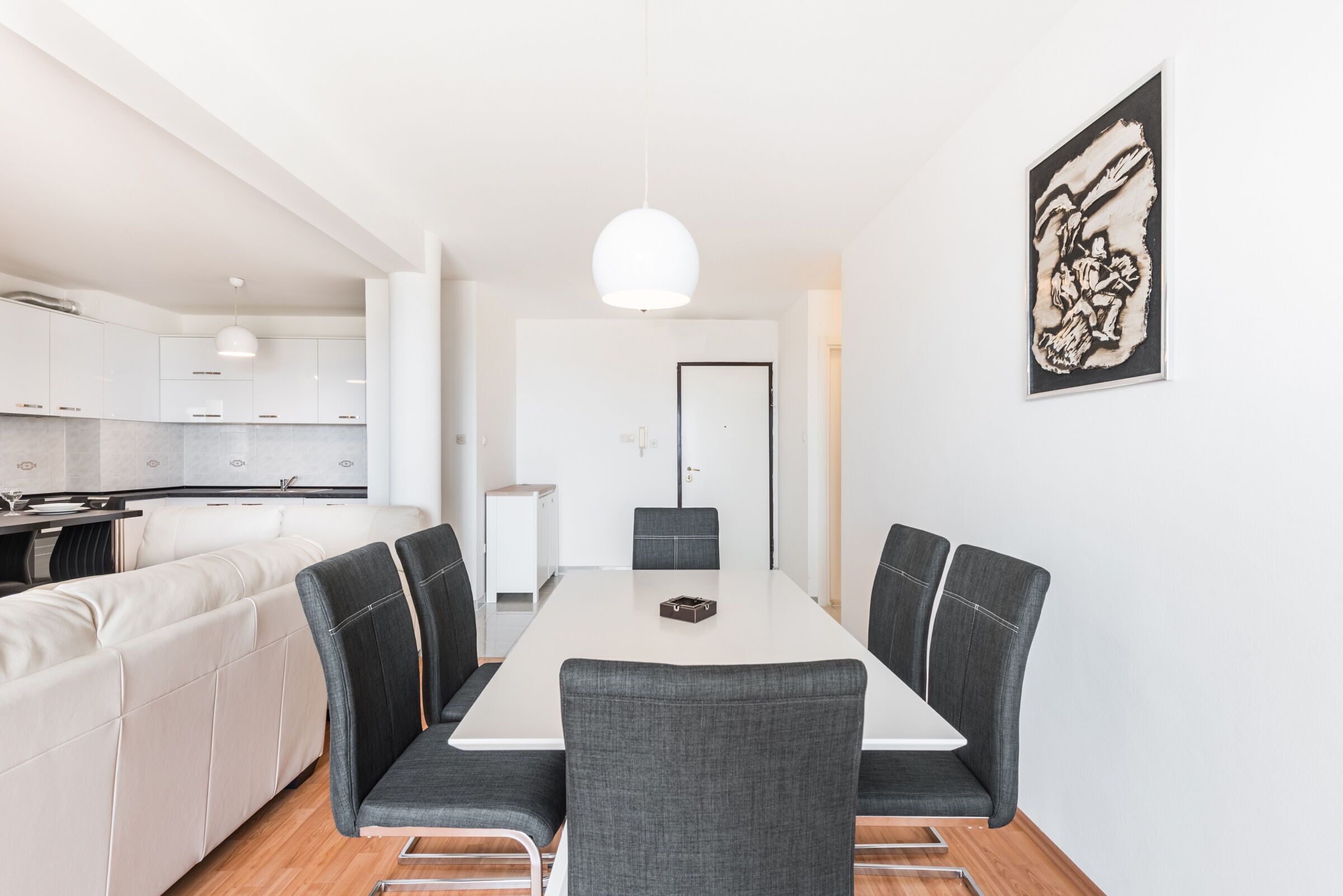 A modern dining area with a white table and grey upholstered chairs, a white sofa, and a kitchen in the background.