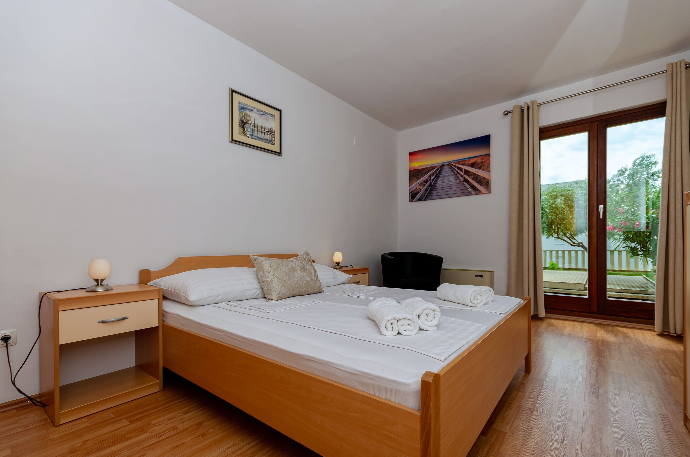 A neatly made bed with white linens and rolled towels, a bedside table with a lamp, a small drawer, and a nightstand. There is a painting on the wall and a window with curtains.
