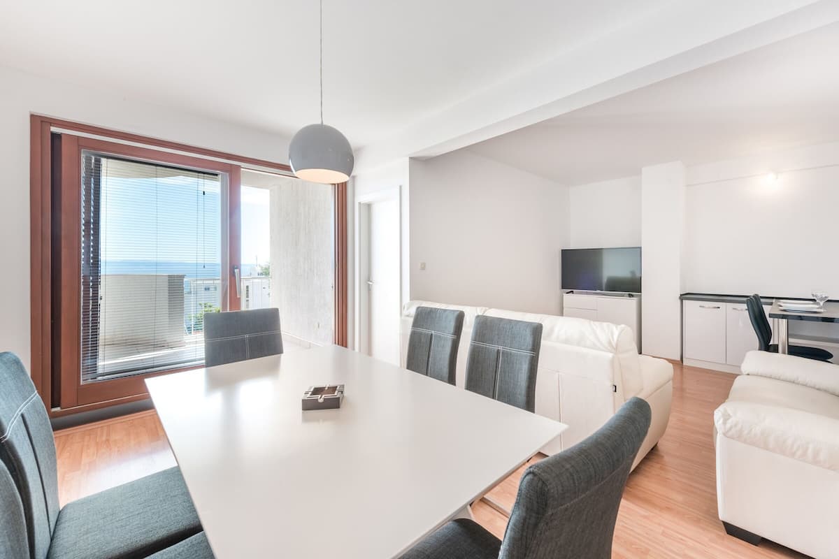 A modern living room with a dining table, grey sofas, and a TV in the background.