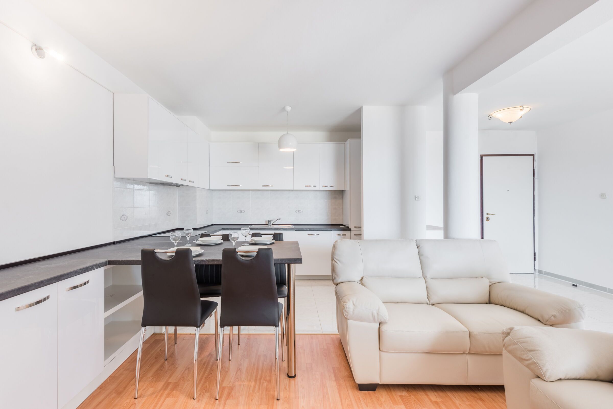 A modern kitchen with white cabinets, a dining area with two chairs, and a beige sofa.
