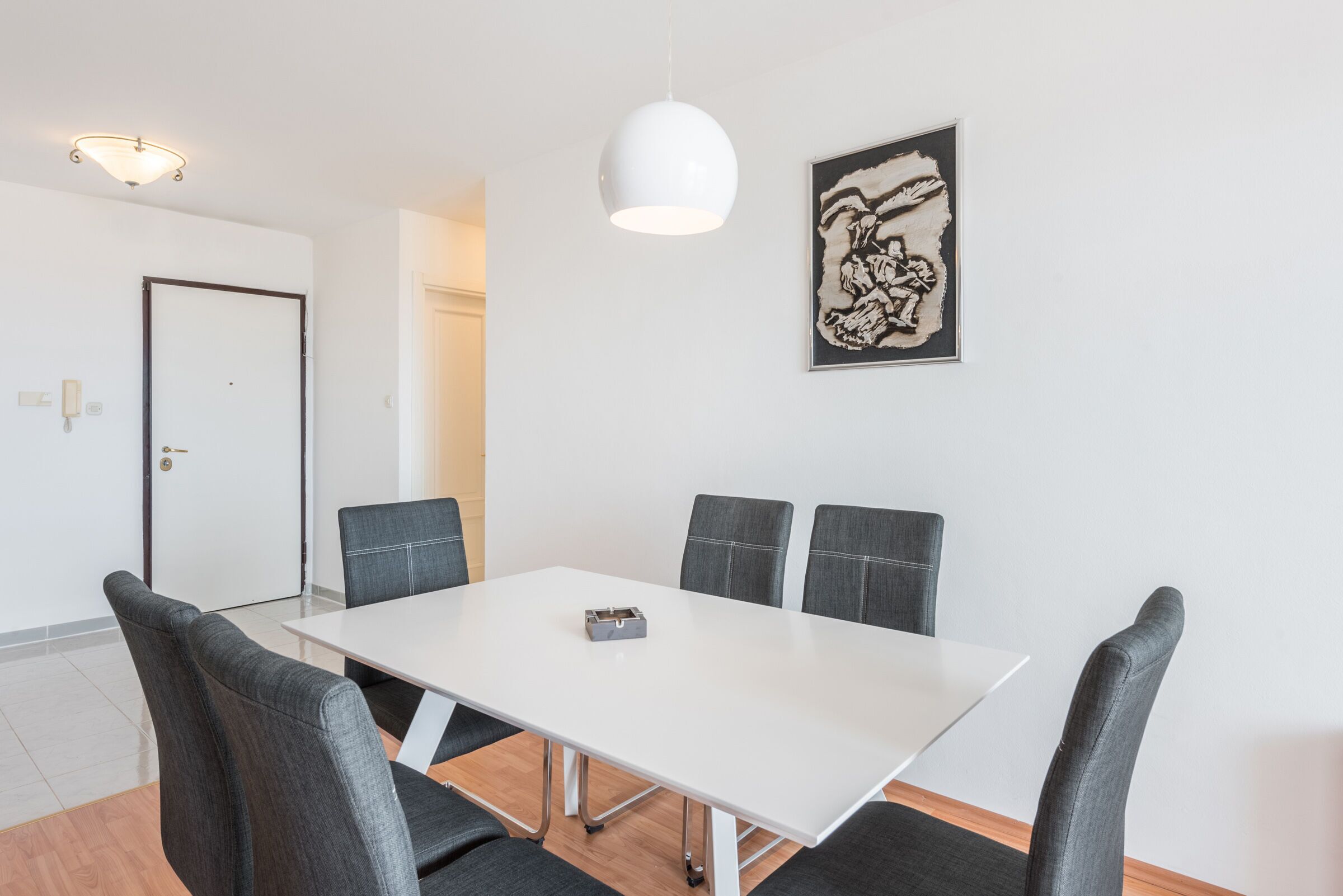 A conference room with a white table, four grey chairs, a white pendant light, and a framed abstract artwork on the wall.