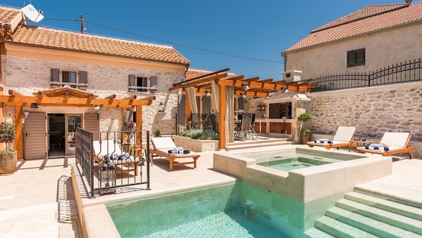 A pool area with lounge chairs and a pergola, surrounded by stone buildings with tiled roofs. - Villa Stone (Skradin)