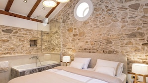 A bedroom with a stone wall, a large bed with white bedding, a round skylight, and a bathtub. - Villa Stone (Skradin)