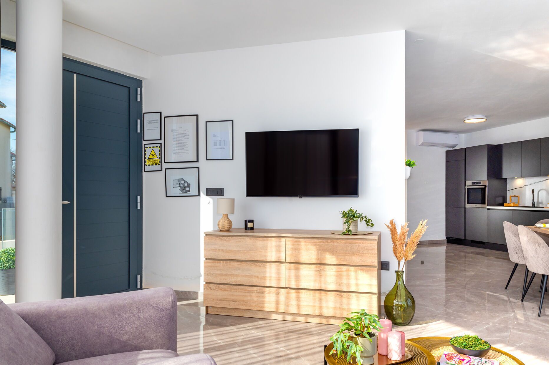 A modern living room with a flat-screen TV, wooden furniture, and a dining area with a kitchen in the background.