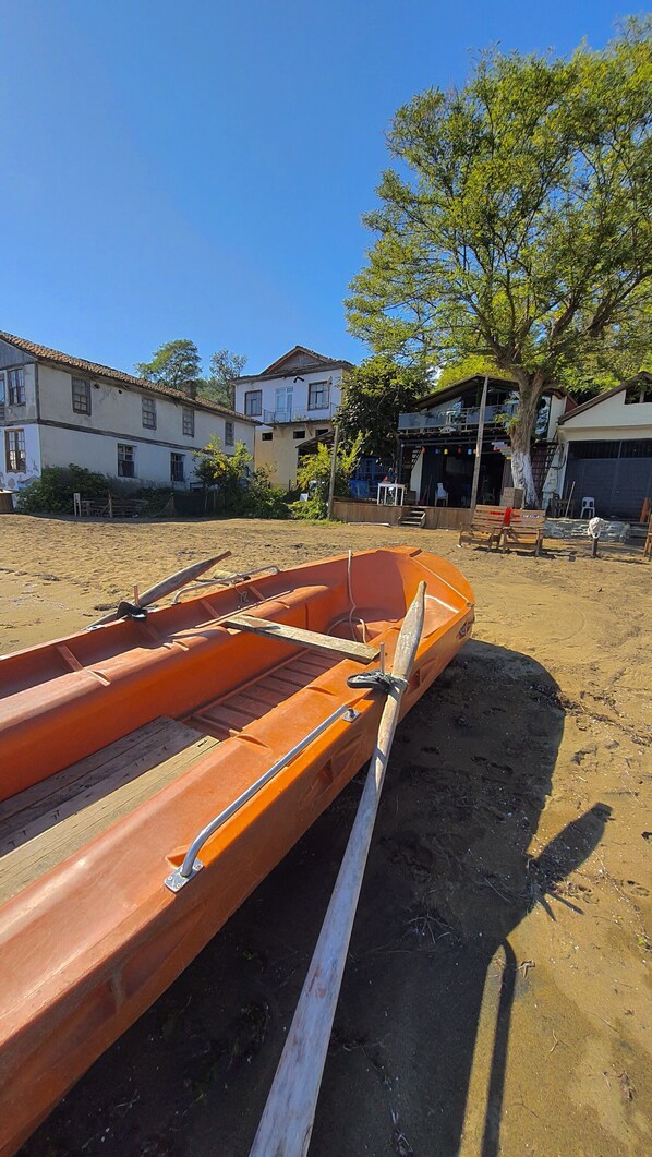 A rowboat tied to a wooden post on a sandy beach with houses and a tree in the background. - Çeşmeönü Bungalov (Persembe)