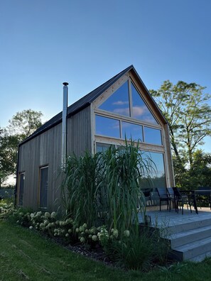 A modern wooden building with large windows and a chimney, surrounded by lush greenery and a patio area. - GREEN LAKESIDE HOUSE   (Pogobie Tylne)