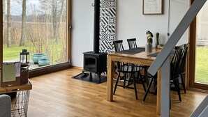 Una habitación amplia con una gran mesa de madera, sillas negras, un estufa de leña negra y vistas al exterior a través de ventanas grandes.
