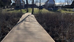 Un paseo marítimo de madera que conduce a una zona residencial con dos casas grandes y cielo despejado.