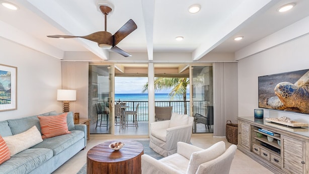 A spacious living room with a balcony overlooking the ocean, a ceiling fan, and a television on a wooden cabinet. - Simply Stunning Condo, C202 is one of of the best! (Lahaina)