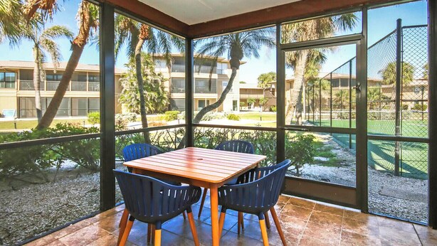 A covered patio with a wooden table and blue chairs, overlooking a landscaped area with palm trees and a building in the background. - Blind Pass E103 - over 1500 sq ft ~ 8 person occupancy (Sanibel)