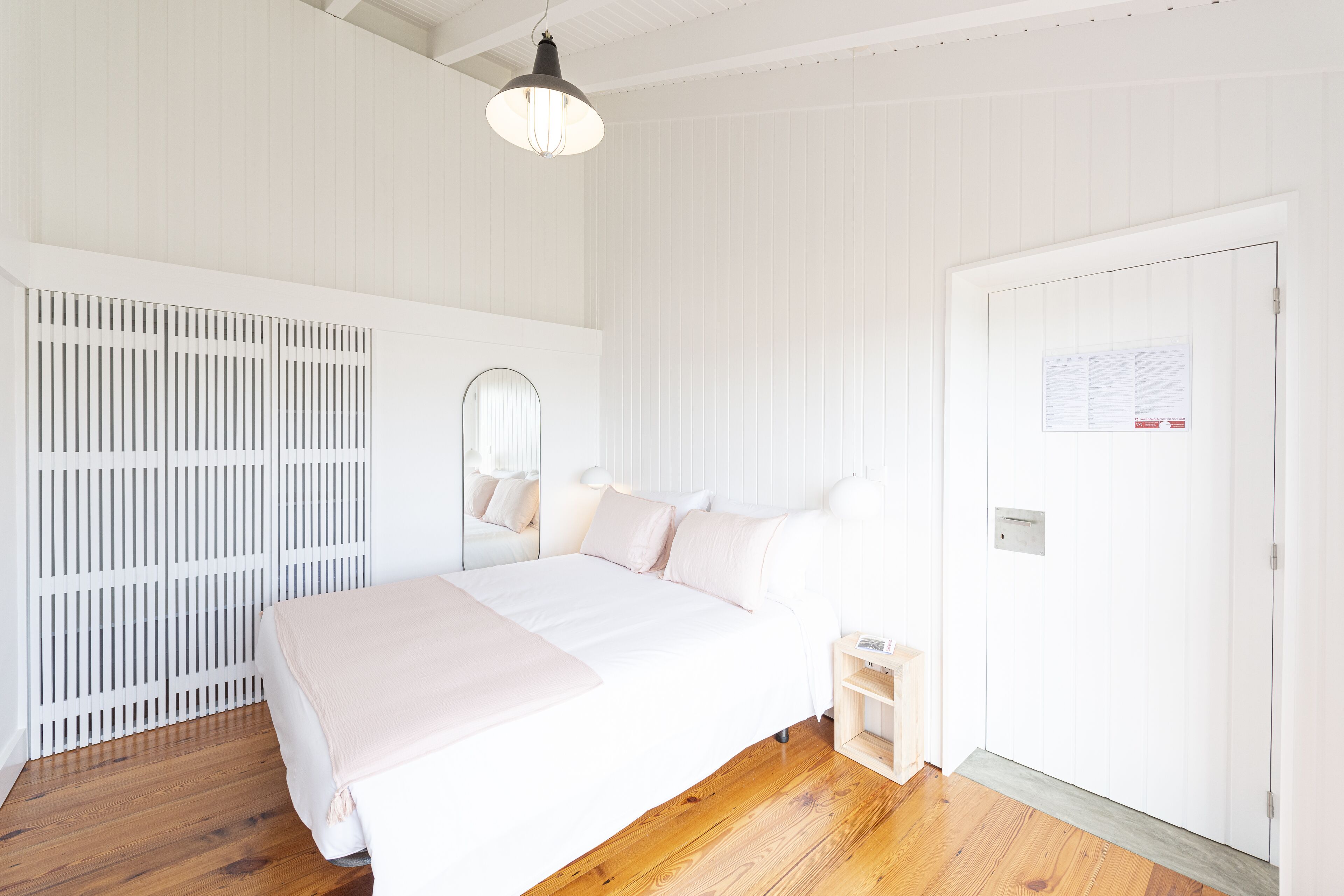 A bedroom with a large bed, a bedside table, a mirror, a wardrobe, and a door with a notice.