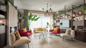 Una sala moderna con dos sofás rojos, un sofá beige y una mesa de centro. Se observa una planta grande, un televisor y estantes con objetos decorativos.