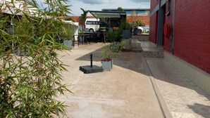 Un espace extérieur avec un bâtiment rouge, un fourgon blanc et un véhicule noir garé devant. On y trouve des plantes et un chemin en béton.