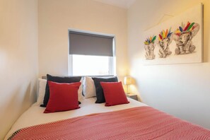 Una camera d'albergo con un letto, cuscini rossi e neri, una coperta a motivi decorativi, una finestra con persiana e un'opera d'arte appesa al muro raffigurante degli elefanti.