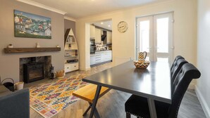 Un espace repas comprenant une grande table, des chaises, une cheminée, un tapis coloré et une vue sur la cuisine.
