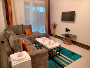 Una sala de estar con un sofá gris, una mesita de centro blanca, un televisor en la pared y una pequeña mesita auxiliar.
