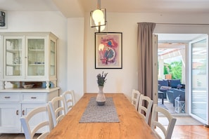 Un comedor con una mesa de madera, sillas blancas, un aparador con puertas de vidrio, un florero con flores y un cuadro en la pared.