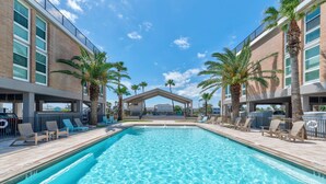 Una piscina con sillones de descanso y palmeras, rodeada de edificios con ventanales amplios y una zona cubierta al fondo.