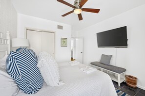 A hotel room with a bed, a television, a ceiling fan, a bench, and a wall-mounted TV.
