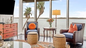 Amplia habitación con un gran ventanal con vistas a una masa de agua, que cuenta con una zona de estar acogedora con sofá y sillones, una mesita con lámpara y un aparador decorativo.