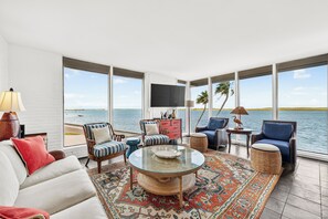 A spacious living area with a large window overlooking a body of water, furnished with a sofa, chairs, and a coffee table.