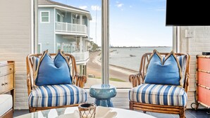 Una habitación con dos sillones de mimbre con cojines azules, una mesita pequeña azul y vista a una playa y una casa al otro lado del agua.