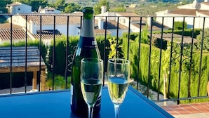 En balkong med en flaska vin och två glas, med utsikt över ett bergigt landskap med hus.