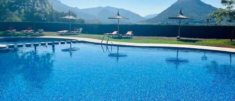A clear blue swimming pool with lounge chairs and umbrellas, surrounded by mountains.