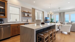 A modern kitchen with a central island, wooden cabinets, and a dining area with a view. - Kamps Baai, Beachfront Home, Golf Cart, 2 Pools (Miramar Beach)