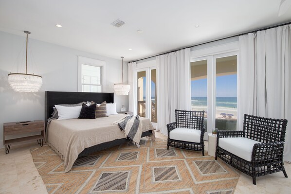 Quarto com cama, duas cadeiras, mesa de cabeceira e vista para a praia.