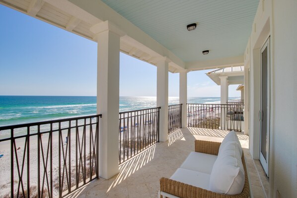 A covered balcony with a view of the ocean, white wicker furniture, and a metal railing.