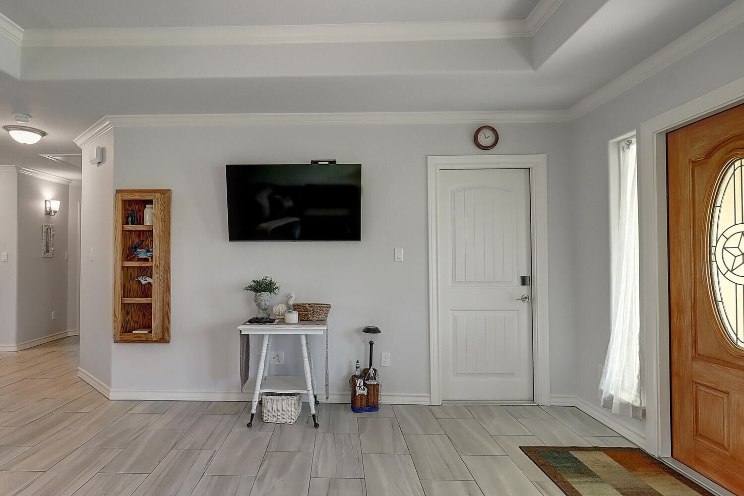 A hallway with a white door, a wooden door with a glass panel, a white wall-mounted TV, a small white table with a vase and a basket, and a wooden bookshelf.