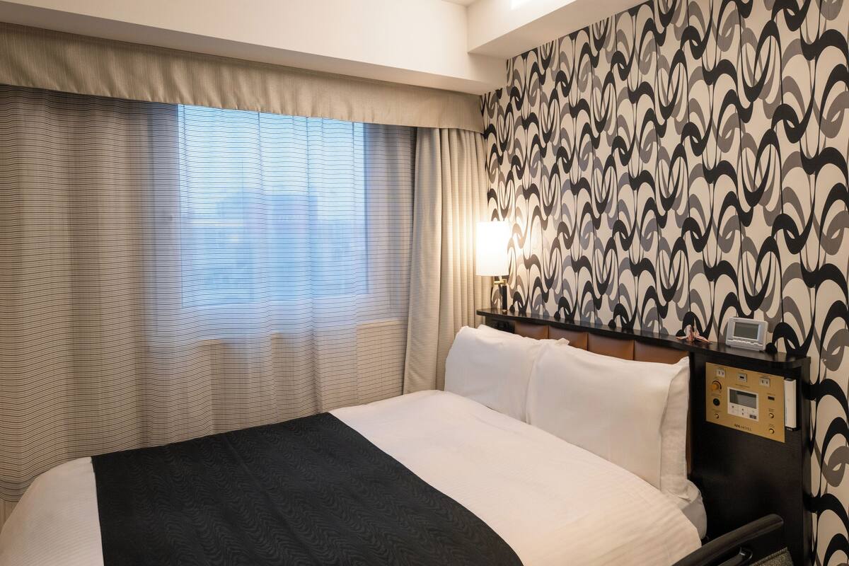 Hotellihuone, jossa on suuri sänky, yöpöytä lampun kanssa ja kuviollinen tapetti seinällä.