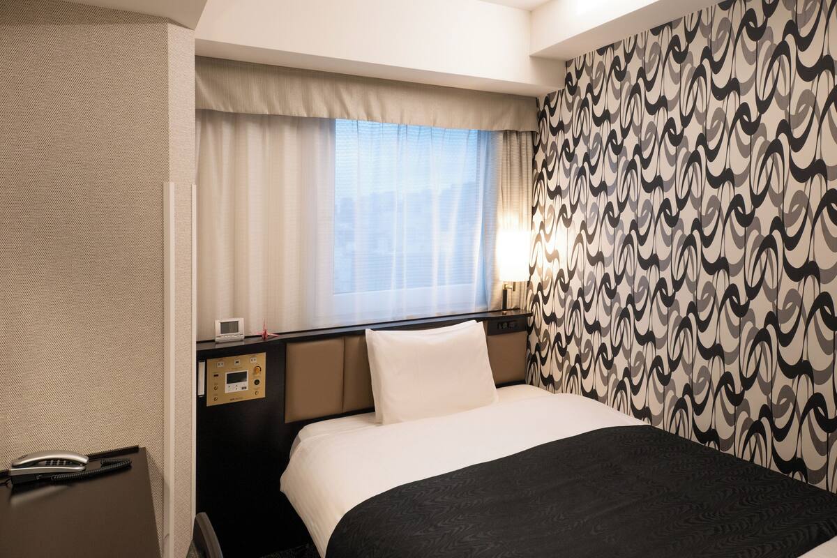 Hotellihuone, jossa on sänky, työpöytä, jossa on puhelin, ohjauspaneeli ja ikkuna, jossa on verhot.